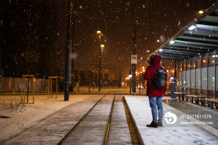 man in red winter coat standing at bus tram stop waiting for public transport