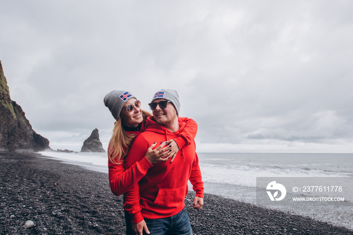 A guy and a girl in red sweaters and gray hats are hugging on a black beach, next to Vic. Happy young couple in a hat with a flag. A happy stylish smiling couple walking and kissing in Iceland.