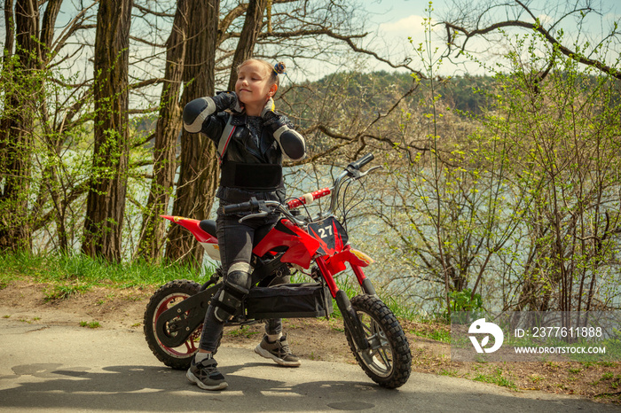 A child girl in motorcycle protection, knee pads and elbow pads on a cross motorcycle stands enjoying a walk on an asphalt path in the park near the river and trees in the spring