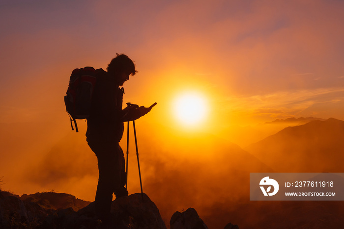 backlit silhouette of hiker with backpack using gps apps on his mobile phone