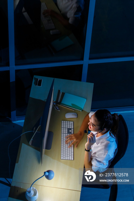 Top view shot of Asian professional female assistant service helpline operator wears headphone microphone working help talking with customer online typing keyboard looking at computer monitor at night