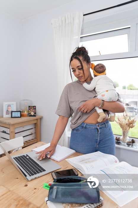 Young mother using laptop while carrying newborn baby girl at home