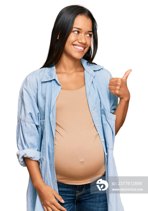 Beautiful hispanic woman expecting a baby showing pregnant belly smiling with happy face looking and pointing to the side with thumb up.