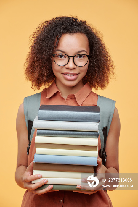 Portrait of smiling clever black girl in eyeglasses holding heap of books while preparing for seminar