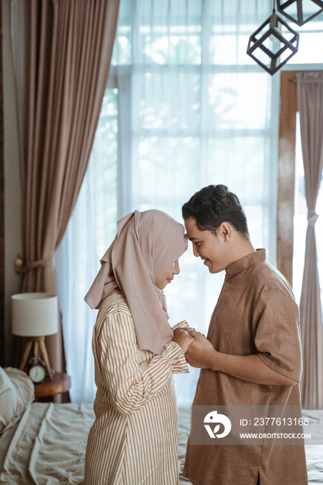 young Muslim couple smiling at each other forehead facing each other and holding hands while standing in the room