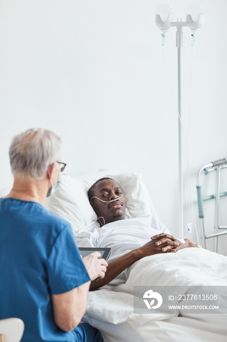 Vertical portrait of young African-American man lying in hospital ed and talking to senior doctor