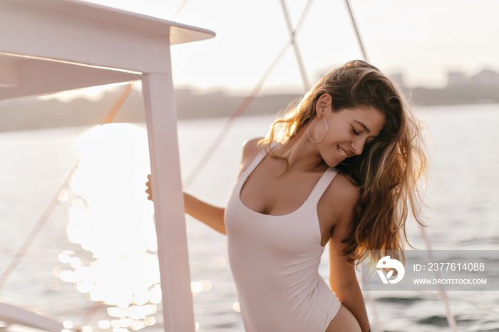 Engaging young blonde with well-built tanned body and sunlit hair in white swimsuit and round earrings posing on yacht against sea background, smiling and looking down