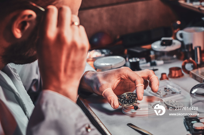 Mature clockmaster is fixing old watch for a customer at his busy repairing workshop.