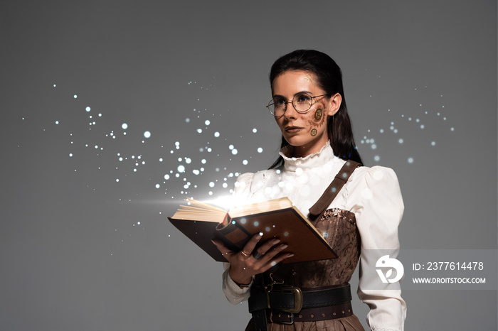 pensive steampunk woman in glasses holding open book with fairy glowing illustration isolated on grey