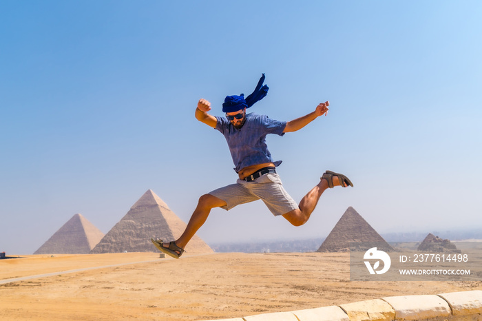 A young tourist jumping for joy in a blue turban and sunglasses at The Pyramids of Giza, the oldest Funerary monument in the world. In the city of Cairo, Egypt