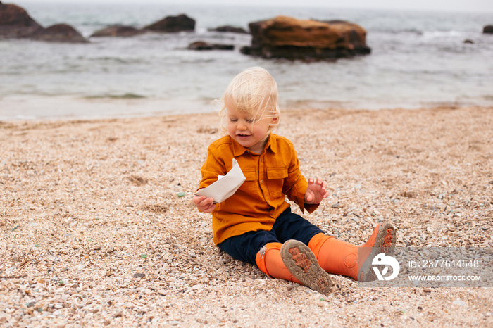 Baby boy is playing with paper boat on the beach near sea. Child on vacation in summer or autumn at the sea on bad weather in autumn or summer time