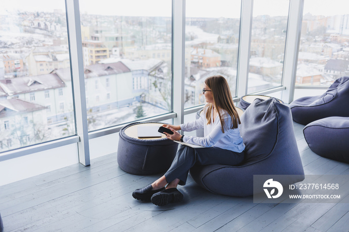 Young female manager in glasses sits on a soft pouf after a working day, modern workspace in the office