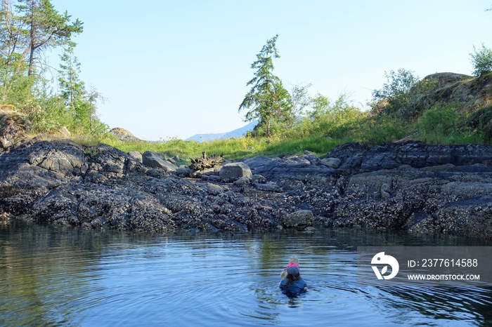 A woman swimming with her dog on a paddleboard along the beautiful rocky shores and calm ocean water of the Harmony Islands, hotham sound, jervis inlet, British Columbia, Canada