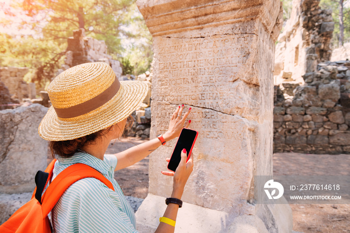 Woman philologist and tourist with smartphone reads and translates ancient Greek from columns in the ruins of an antique city. Linguistics and archaeology