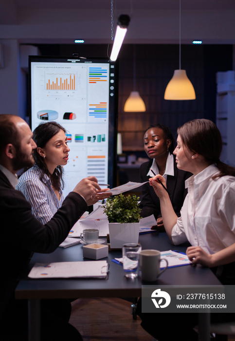 Businesswoman leder sitting at desk analyzing marketing presentation discussing financial graps paperwork. Diverse multi-ethnic teamwork brainstorming project ideas working in meeting room late night