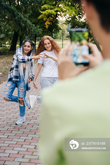 Rear view of a boy filming his girlfriends copying a trend from the phone app, showing the heart sign. Boy filming two girls. Selfie creative idea. Streaming