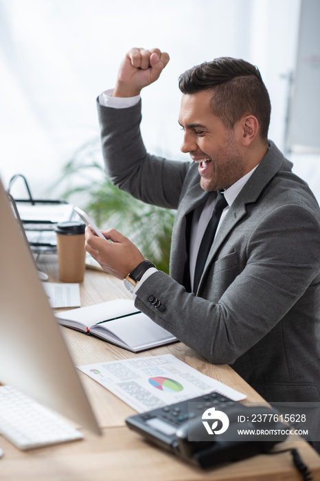 excited trader showing win gesture while messaging on smartphone on blurred foreground