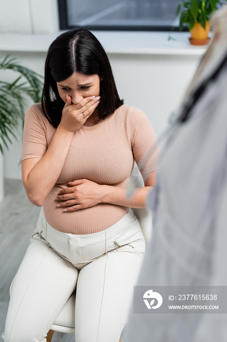 upset pregnant woman covering mouth with hand near blurred doctor in consulting room