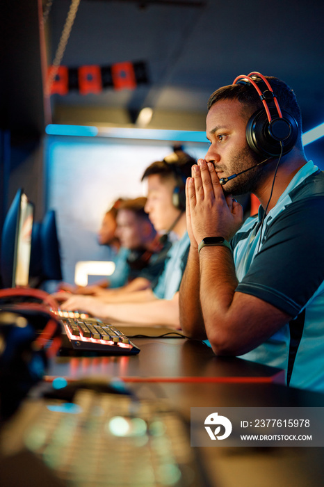 Serious young man in headset focused on video game while sitting at modern computer in internet cafe