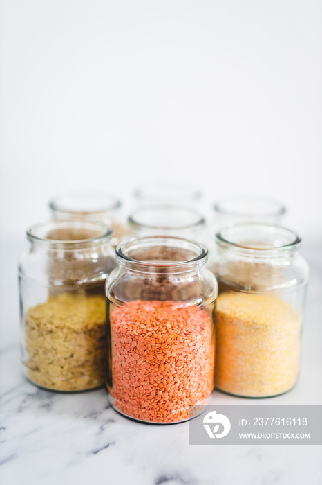 healthy whole grains and legumes in clear pantry jars on marble background including quinoa rice buckwheat lentils and barley, simple ingredients concept