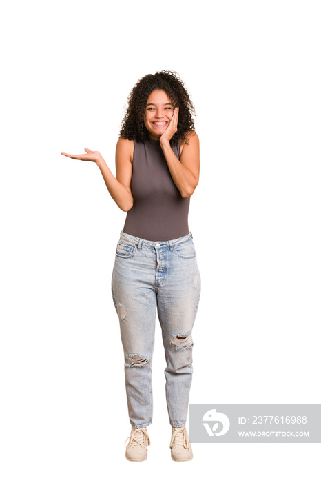 Young african american woman with curly hair cut out isolated holds copy space on a palm, keep hand over cheek. Amazed and delighted.