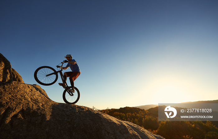 Silhouette of professional cyclist riding on back wheel on trial bicycle. Sportsman rider making trick on the edge of rock on the top of mountain at sunset. Concept of extreme sport active lifestyle