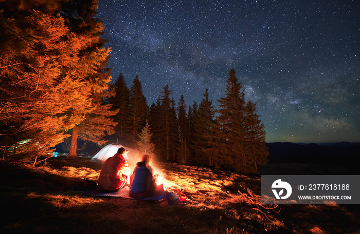 Night camping near forest. Couple of tourists spending romantic evening outdoors by bonfire on lawn. On the background tents, pines, starry sky. Active recreation in nature. Motion blur.