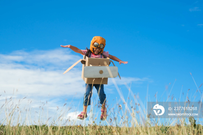 Cute dreamer little girl playing with cardboard planes in the meadow on a sunny day. Happy kid playing with cardboard plane against blue summer sky background. Childhood dream imagination concept.