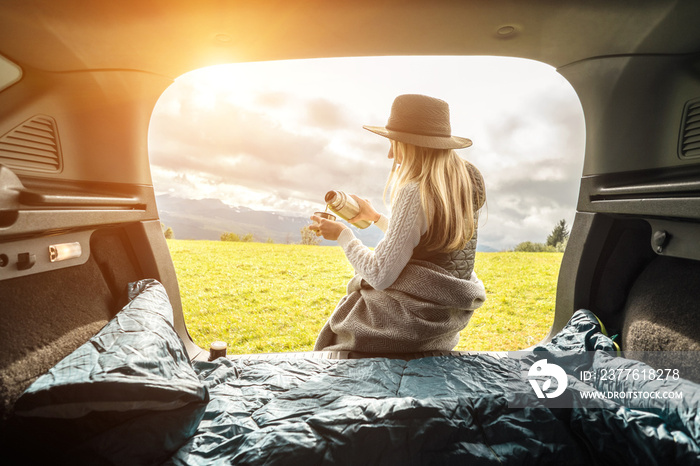 Girl resting in her car. Woman hiker, hiking backpacker traveler camper in sleeping bag, drinking hot tea and relaxing on top of mountain. Health care, authenticity, sense of balance and calmness.