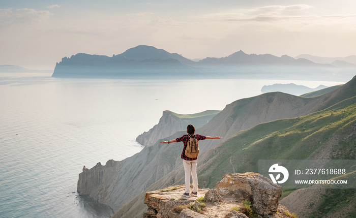 A young, happy woman with a backpack standing on a rock with her hands up. Mountain and coastal travel, freedom and an active lifestyle. Breathtaking scenery of the Black Sea coast