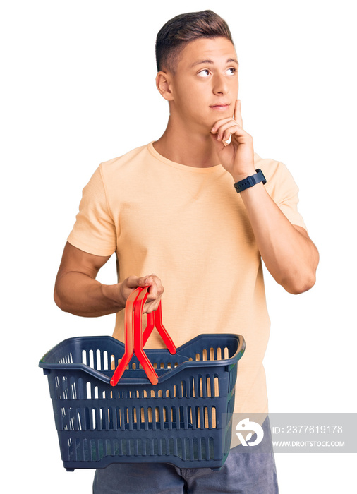 Young handsome hispanic man holding supermarket shopping basket serious face thinking about question with hand on chin, thoughtful about confusing idea