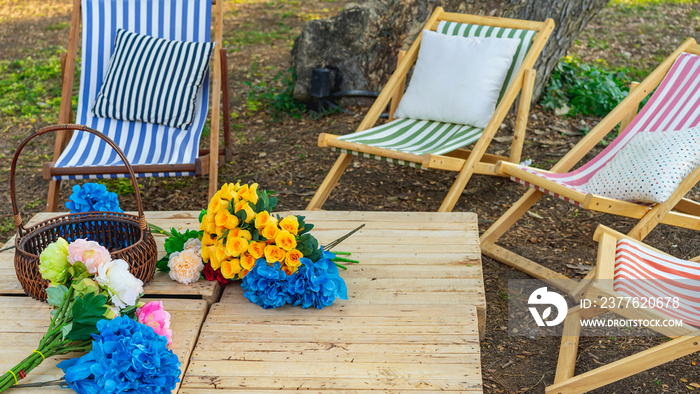 Beautiful artificial bouquets of various colors placed on wooden table among deck chairs in garden. Multi-colored artificial flowers adorn the wooden table for beautiful and relaxing. Selective focus.