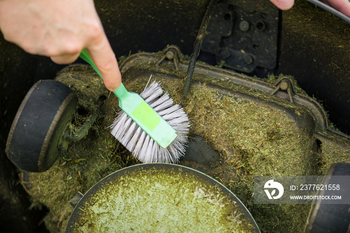 Close up view of man hand brushing off layer of wet grass stuck under automated lawnmower, maintenance concept.