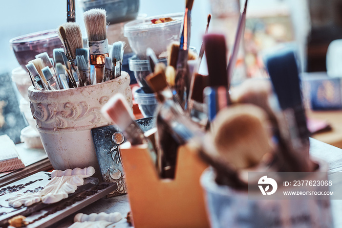 A lot of different brushes on artist’s table in jars. There are artistick’s mess on the table.