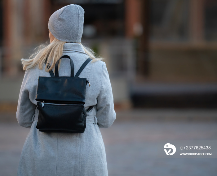 Happy young blonde teenage girl going to school or college wearing black backpack. Blonde girl in rgay hat and gray coat. Pictuce from back