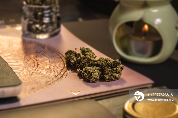 Cannabis buds on notebooks with glass jar, gold lid, incense burner, tealight and black background