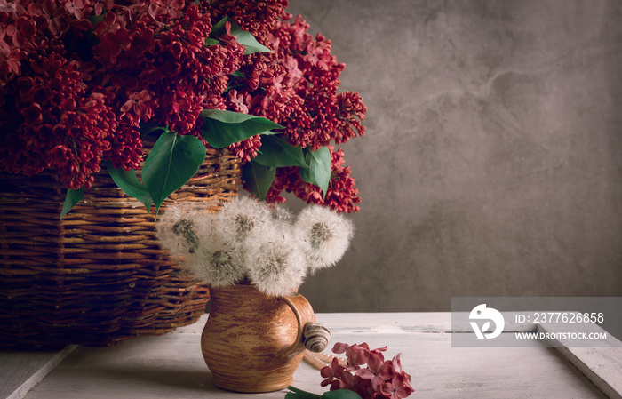 Beautiful dandelion bouquet in a brown ceramic pot against purple lilac flowers in dark moody style. Vintage filtered photo.