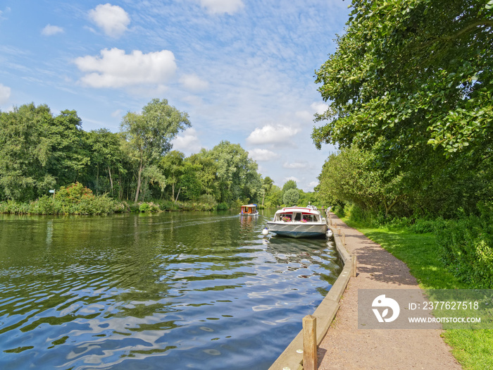 A large cabin cruiser moored on the banks of the River Bure, on the Norfolk Broads near Wroxham, while a second boat travels upstream.