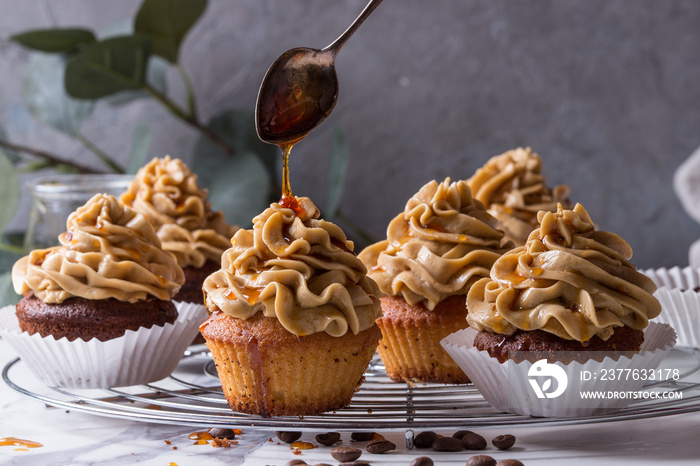 Fresh baked homemade cupcakes with coffee buttercream and pouring from spoon caramel standing on cooling rack with eucalyptus branch and coffee beans above over white marble kitchen table.