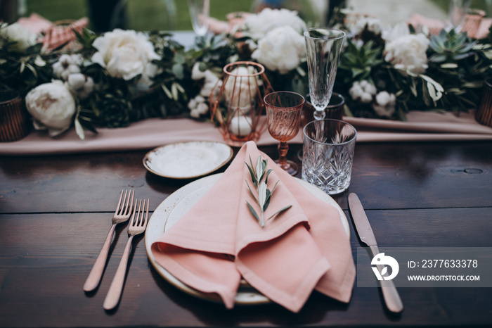 on wooden long banquet table are glasses, plates, candles, table is decorated with compositions of greenery and eucalyptus branches, plates are decorated with napkins and sprig of Italian greenery