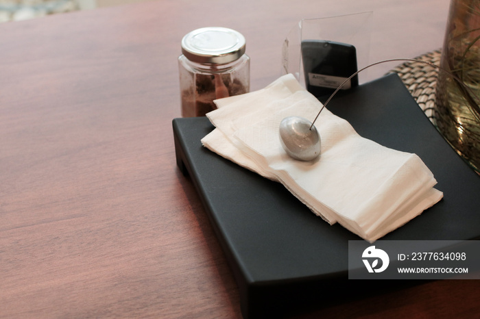 Metal paperweight on stack of white square paper napkin. modern paper napkin holder Close up napkin holder with paperweight to keep brown napkins on black plate and wooden table in coffee shop