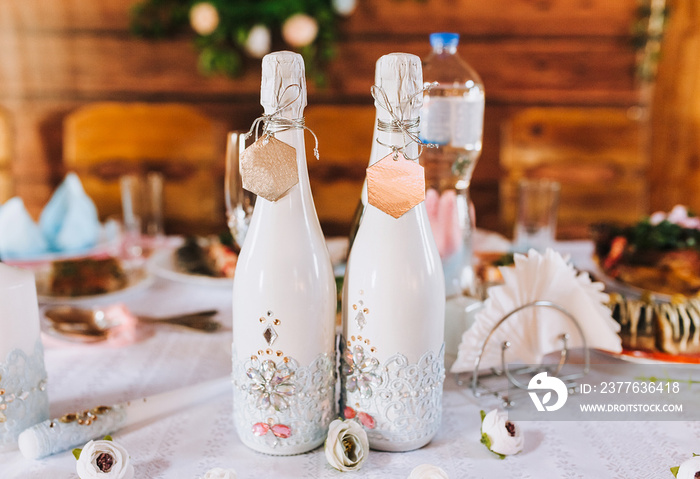 A white bottle of champagne, decorated with precious stones, jewelry with a metal, gold tag, tied with a thread stands on a wooden table with candles. Wedding photography, holiday decorations.