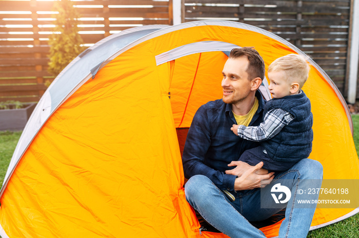 Family day. Father and little son sit in tent in the backyard, they play camp