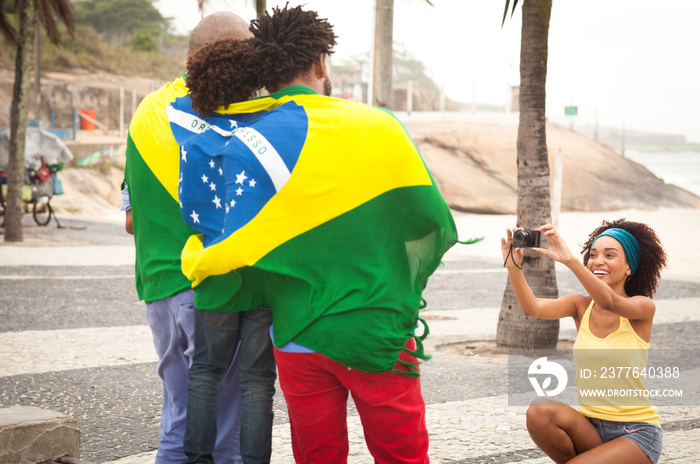 Young mother photographing family wrapped in Brazilian flag on Ipanema beach, Rio De Janeiro, Brazil