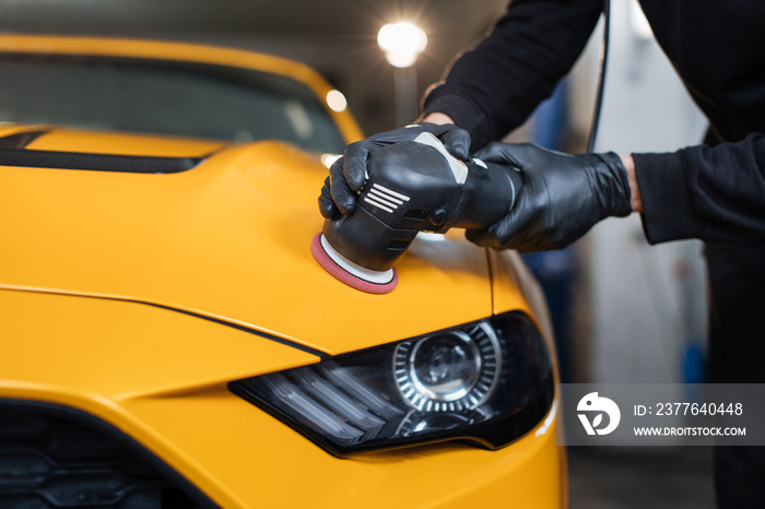 Car detailing and polishing concept. Hands of professional car service male worker, with orbital polisher, polishing yellow luxury car hood in auto repair shop.