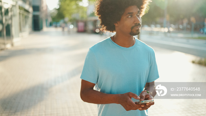 Closeup portrait of  young African American man in light blue t-shirt using his smartphone. Man looks at photos, videos in his mobile phone.