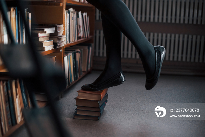 Standing on books and trying to reach top of shelves. Female student is in library. Conception of education