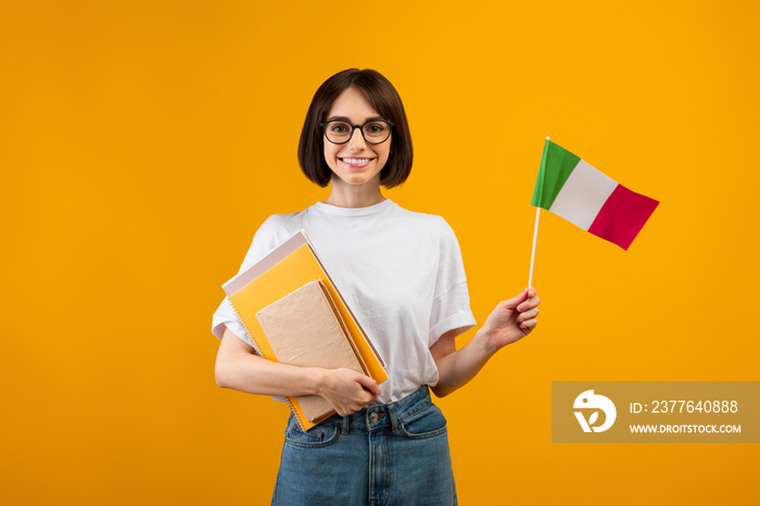 Learning Italian, student exchange and travel. Happy lady holding notebooks and small flag of Italy, orange background