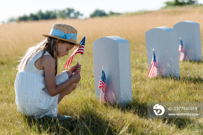 sad kid in straw hat sitting near headstones and holding american flag