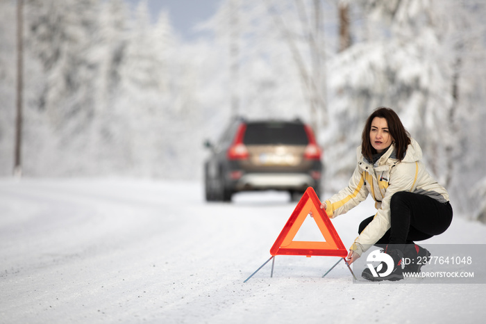 Pretty, young woman setting up a warning triangle and calling for assistance after her car broke down in the middle of nowhere on a freezing winter day